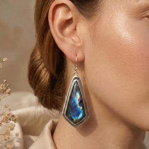 Puede incluir: Un pendiente plateado con forma geométrica de diamante alargado. El pendiente presenta una vibrante piedra azul y dorada iridiscente engastada en un marco plateado. El pendiente cuelga de un sencillo gancho plateado.