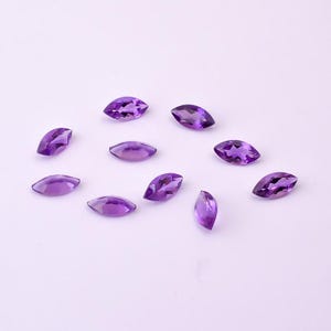May include: A collection of marquise-cut amethyst gemstones, each displaying a vibrant purple hue. The gemstones are scattered across a white surface, showcasing their polished facets and unique shape. Ideal for jewelry making or gemstone collecting.