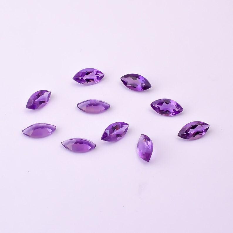 May include: A collection of marquise-cut amethyst gemstones, each displaying a vibrant purple hue. The gemstones are scattered across a white surface, showcasing their polished facets and unique shape. Ideal for jewelry making or gemstone collecting.