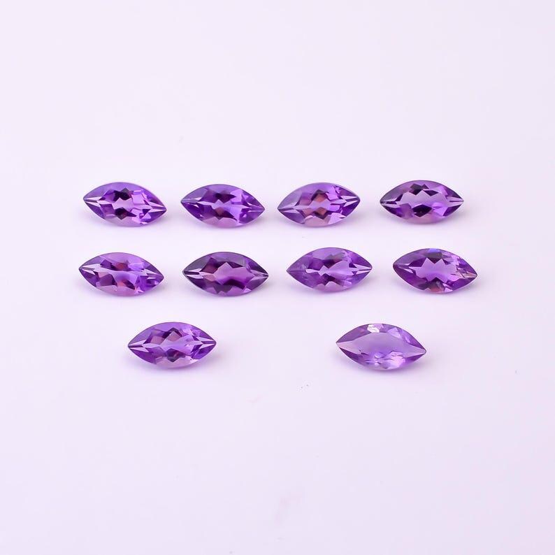 May include: A collection of eleven marquise-cut amethyst gemstones. The gemstones are a deep purple color and are arranged on a white surface. The gemstones are faceted, with a pointed tip on each end.