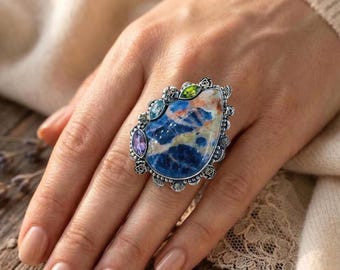 Könnte beinhalten: Ein auffälliger Ring mit einem großen, tropfenförmigen blauen Edelstein mit weißen und goldenen Adern. Der Ring ist mit kleineren Edelsteinen in Lila, Grün und Blau besetzt, die in einem silberfarbenen Metall mit detailliertem floralem Design gefasst sind. Ein einzigartiges Schmuckstück.