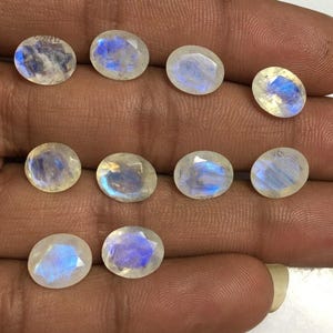 May include: A collection of oval-shaped moonstone gemstones. Each stone displays a milky white base with iridescent blue and white flashes. The gemstones are faceted, catching light to create a shimmering effect. The stones are displayed on a hand.