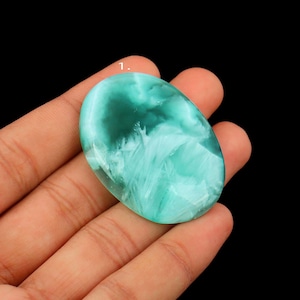 May include: An oval-shaped gemstone with a swirling pattern of teal and white. The polished stone is held in a hand against a black background. The gemstone's surface has a translucent quality.