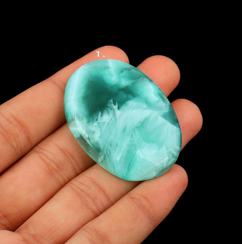 May include: An oval-shaped gemstone with a swirling pattern of teal and white. The polished stone is held in a hand against a black background. The gemstone's surface has a translucent quality.
