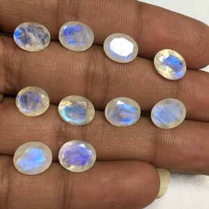 May include: A collection of oval-shaped moonstone gemstones. The stones have a translucent, milky white base with iridescent blue flashes. The gemstones are faceted, catching the light and displaying a subtle shimmer.