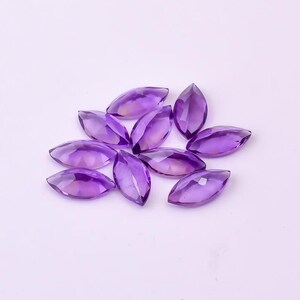 May include: A collection of marquise-cut amethyst gemstones. The gemstones are a deep purple color with faceted surfaces, catching the light. They are arranged on a light purple background.