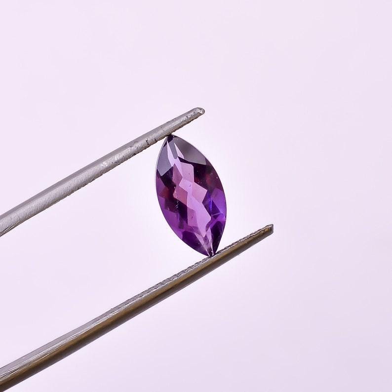 May include: A marquise-cut, faceted amethyst gemstone held by tweezers. The gemstone is a deep purple color with visible internal facets. The tweezers are silver-colored and the background is a soft white.