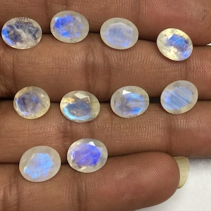 May include: A collection of oval-shaped moonstone gemstones. The stones have a translucent, milky white base with iridescent blue and purple flashes. The gemstones are displayed on a hand, showcasing their polished surfaces and natural beauty.