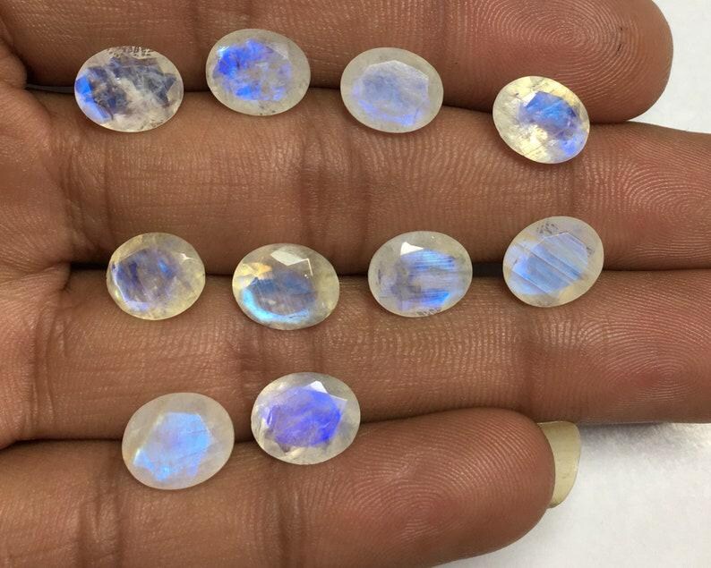 May include: A collection of oval-shaped moonstone gemstones. The stones have a translucent, milky white base with iridescent blue and purple flashes. The gemstones are displayed on a hand, showcasing their polished surfaces and natural beauty.