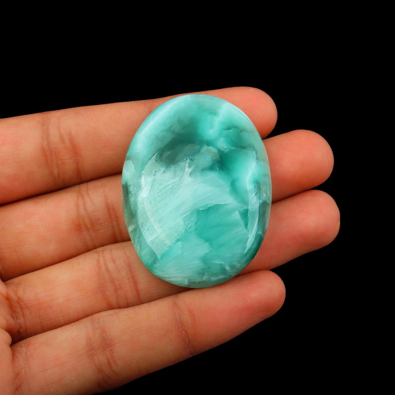 May include: An oval-shaped, polished gemstone with a swirling pattern of light and dark teal and white. The stone has a glossy, reflective surface and is set against a black background.