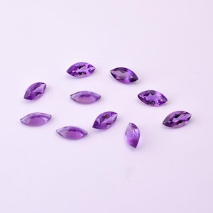 May include: A collection of marquise-cut amethyst gemstones in varying shades of purple. The gemstones are scattered across a light purple surface, showcasing their polished facets and natural color variations. Ideal for jewelry making or gemstone collections.