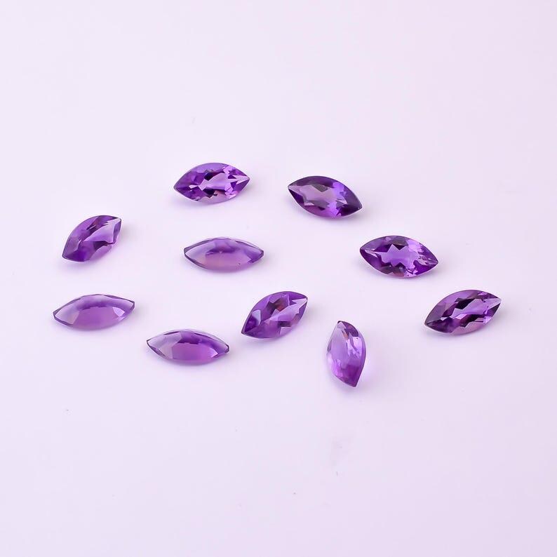 May include: A collection of marquise-cut amethyst gemstones in varying shades of purple. The gemstones are scattered across a light purple surface, showcasing their polished facets and natural color variations. Ideal for jewelry making or gemstone collections.
