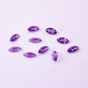May include: A collection of marquise-cut amethyst gemstones in varying shades of purple. The gemstones are scattered across a light purple surface, showcasing their polished facets and natural color variations. Ideal for jewelry making or gemstone collections.
