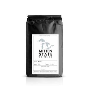 May include: A black coffee bag with a white label featuring the text "MITTEN STATE COFFEE CO." and a map outline. The label also indicates "Blonde" origin, "Light" roast, and "Standard" grind. The bag is filled with fresh roasted coffee.