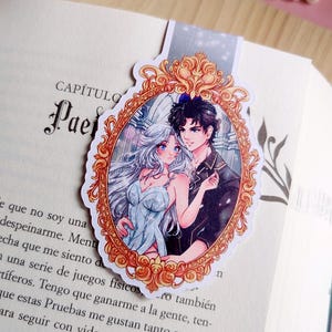 Puede incluir: Marcap&aacute;ginas con una ilustraci&oacute;n de estilo anime de una pareja en un marco dorado ornamentado. La mujer lleva un vestido azul claro y el hombre una chaqueta negra. El marcap&aacute;ginas est&aacute; en un libro abierto, tema rom&aacute;ntico.