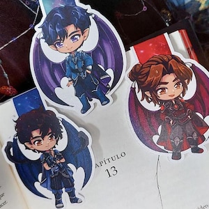 Może przedstawiać: Trzy zakładki do książek z postaciami w stylu anime ze skrzydłami nietoperza umieszczone są na otwartej książce. Postacie mają ciemne włosy i noszą szczegółowe stroje w odcieniach niebieskiego i czerwonego. Strony książki są białe, a tekst "APÍTULO 13" jest widoczny.