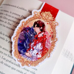 May include: A bookmark featuring a romantic anime-style illustration of a couple embracing within an ornate gold frame. The bookmark is red at the top and is placed in an open book.