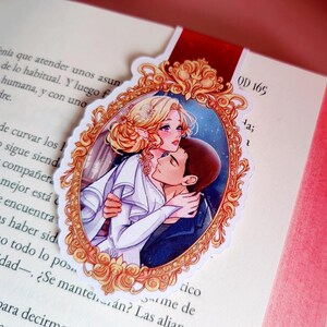 May include: A bookmark featuring a romantic illustration of a couple embracing within an ornate, gold-colored frame. The bookmark is placed on the pages of a book, with text visible. The bookmark has a red ribbon.