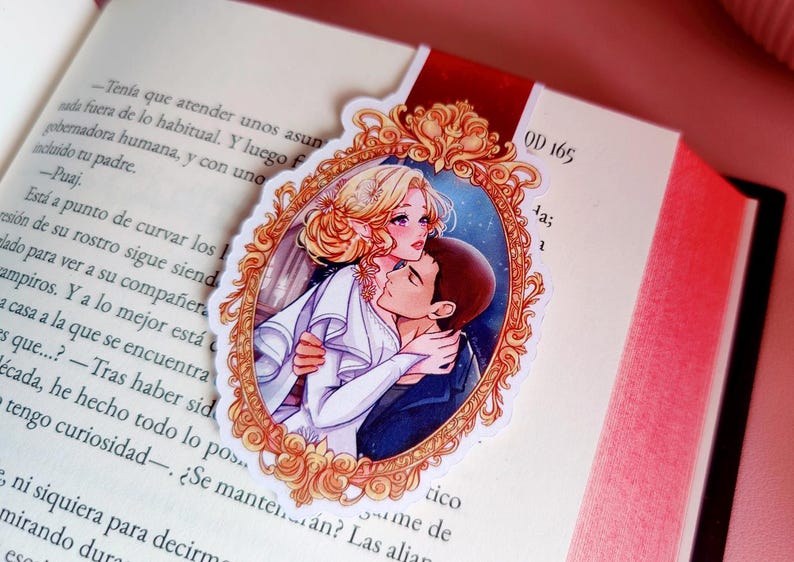 May include: A bookmark featuring a romantic illustration of a couple embracing within an ornate, gold-colored frame. The bookmark is placed on the pages of a book, with text visible. The bookmark has a red ribbon.