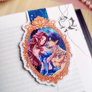 May include: A bookmark featuring a romantic illustration of two fantasy characters within an ornate gold frame. The characters have red and blue hair, and the background depicts a starry night sky. The bookmark is placed on an open book.