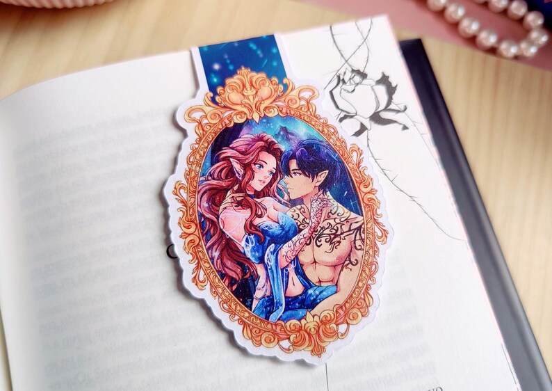 May include: A bookmark featuring a romantic illustration of two fantasy characters within an ornate gold frame. The characters have red and blue hair, and the background depicts a starry night sky. The bookmark is placed on an open book.