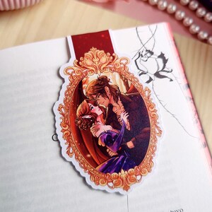 May include: A bookmark featuring a romantic illustration of a couple embracing within an ornate, gold-colored frame. The bookmark is placed on the pages of an open book. The illustration depicts a man and woman in a passionate embrace, with the woman in a purple dress.