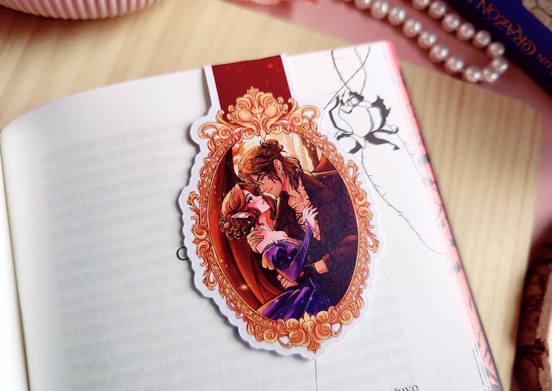 May include: A bookmark featuring a romantic illustration of a couple embracing within an ornate, gold-colored frame. The bookmark is placed on the pages of an open book. The illustration depicts a man and woman in a passionate embrace, with the woman in a purple dress.
