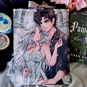 Puede incluir: Una funda de libro con una ilustración de estilo anime de una pareja. La funda se combina con un libro titulado "Powerless" de Lauren Roberts. También se ven una vela y un tarro con tapa.