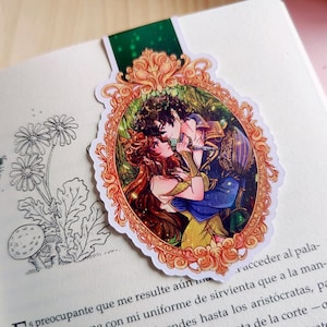 Puede incluir: Un marcap&aacute;ginas con una ilustraci&oacute;n rom&aacute;ntica de una pareja abraz&aacute;ndose dentro de un marco dorado ornamentado. La obra de arte representa a un hombre y una mujer con atuendos coloridos, sobre un fondo exuberante. El marcap&aacute;ginas est&aacute; en un libro abierto.