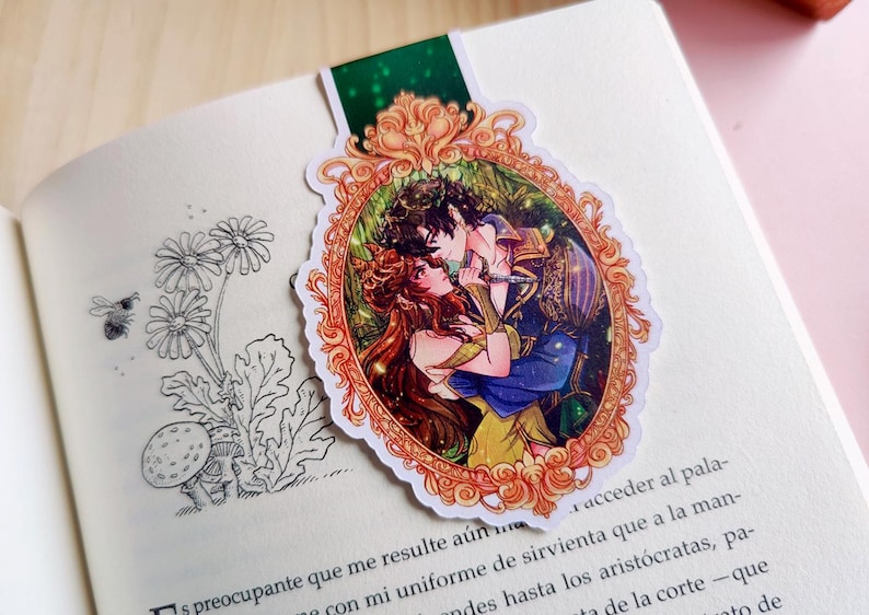 Puede incluir: Un marcap&aacute;ginas con una ilustraci&oacute;n rom&aacute;ntica de una pareja abraz&aacute;ndose dentro de un marco dorado ornamentado. La obra de arte representa a un hombre y una mujer con atuendos coloridos, sobre un fondo exuberante. El marcap&aacute;ginas est&aacute; en un libro abierto.