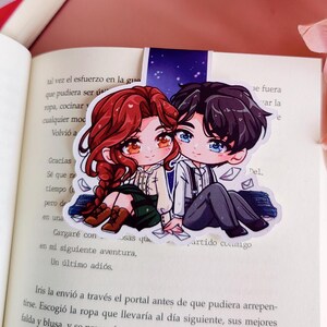 May include: A bookmark depicting a cartoon couple, seated together. The woman has red hair, and the man has black hair. The bookmark is inserted in an open book with Spanish text. The background features a starry night design.
