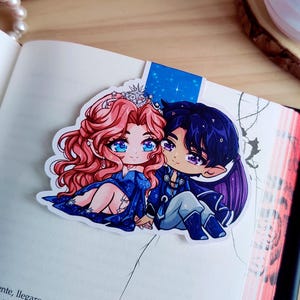 Può includere: Un segnalibro con una coppia in stile anime. Il personaggio femminile ha i capelli rosa e una corona, mentre il personaggio maschile ha i capelli scuri. Indossano abiti blu e si tengono per mano. Il segnalibro è su un libro aperto.