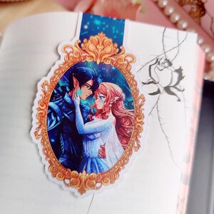 Puede incluir: Un marcap&aacute;ginas con una ilustraci&oacute;n rom&aacute;ntica de una pareja abraz&aacute;ndose dentro de un marco dorado ornamentado. La obra de arte representa a un hombre con cabello oscuro y una mujer con cabello rojo, sobre un fondo azul y turquesa.