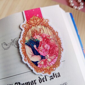 May include: A bookmark featuring a romantic illustration of a couple kissing, set within an ornate gold frame. The bookmark is placed on an open book with text visible. The top of the bookmark is pink.