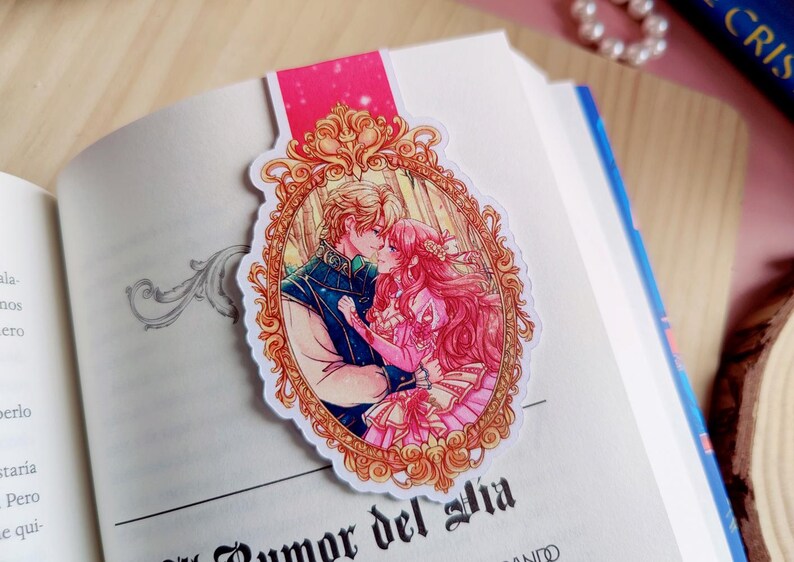 May include: A bookmark featuring a romantic illustration of a couple kissing, set within an ornate gold frame. The bookmark is placed on an open book with text visible. The top of the bookmark is pink.
