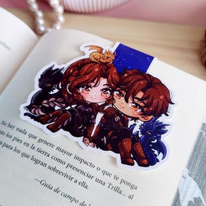 May include: A colorful anime-style bookmark featuring two characters with brown hair and matching outfits, accompanied by small dragon figures. The bookmark is placed on an open book.