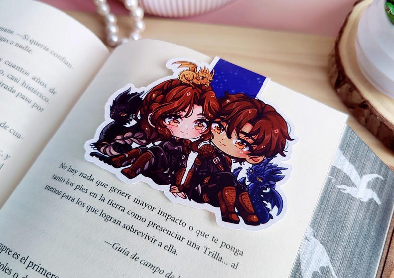 May include: A colorful anime-style bookmark featuring two characters with brown hair and matching outfits, accompanied by small dragon figures. The bookmark is placed on an open book.