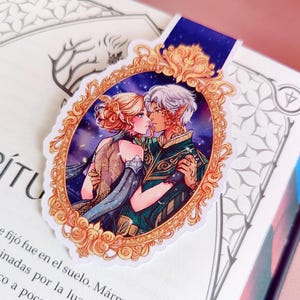 May include: A decorative bookmark featuring a romantic illustration of two figures embracing, set within an ornate gold frame. The image has a starry background and a blue ribbon. The bookmark is placed on an open book.