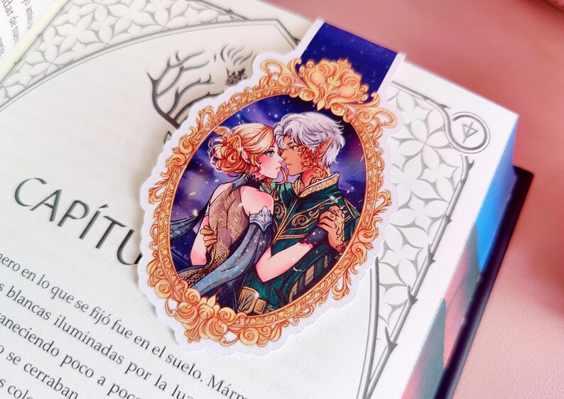 May include: A decorative bookmark featuring a romantic illustration of two figures embracing, set within an ornate gold frame. The image has a starry background and a blue ribbon. The bookmark is placed on an open book.