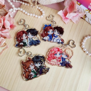 May include: Four acrylic keychains featuring cartoon couples with heart-shaped clasps. Each keychain depicts a pair of characters with different hair colors and outfits. The keychains are arranged on a light wooden surface, with pink flowers and pearl necklaces in the background.