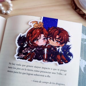 Puede incluir: Un marcapáginas colorido con dos personajes de estilo anime con cabello rojo y marrón, cada uno con un dragón. El marcapáginas está en un libro abierto con texto en español. Un pequeño dragón dorado está en la cabeza de un personaje.