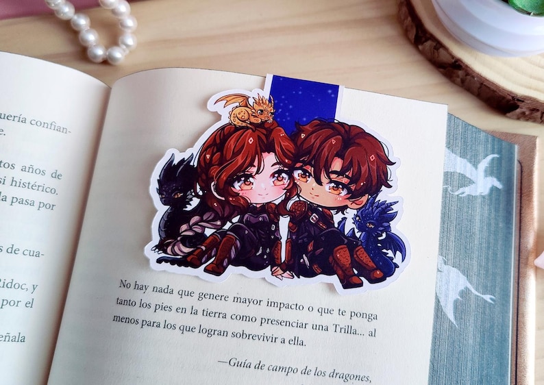May include: A colorful bookmark featuring two anime-style characters with red and brown hair, each with a dragon companion. The bookmark is placed on an open book with text in Spanish. A small gold dragon sits atop one character's head.