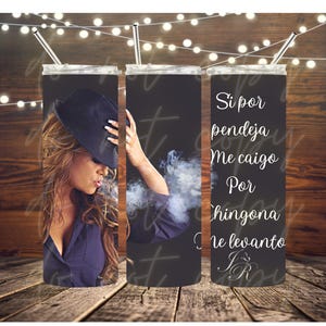 May include: A black tumbler with a woman wearing a fedora and a navy blue shirt. The tumbler has the text "Si por pendeja Me caigo Por chingona Me levanto" in white script. A silver straw is inserted in the lid.
