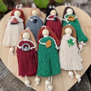 Könnte beinhalten: Eine Sammlung handgefertigter Makramee-Puppen mit Holzköpfen und Kleidern aus Garn in verschiedenen Farben. Jede Puppe hat einzigartige Details, darunter kleine Anhänger und Accessoires. Die Puppen sind auf einem herzförmigen Holzbrett angeordnet.