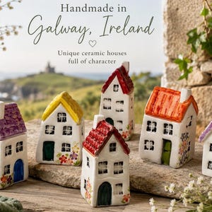 Könnte beinhalten: Sechs kleine Keramikhäuser mit weißen Wänden und farbigen Dächern in Violett, Gelb, Rot und Orange. Jedes Haus hat bemalte Fenster, Türen und florale Details an der Basis. Der Text lautet: Handmade in Galway, Ireland.