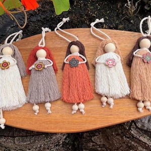 Puede incluir: Cinco adornos de muñecas hechos a mano con cabezas de madera, pelo de hilo y vestidos con flecos en blanco, gris, naranja y beige. Cada muñeca tiene un acento de flor de madera y piernas colgantes de cuentas de madera.