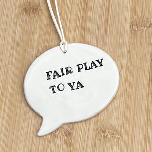 Puede incluir: Un adorno de cerámica blanca con forma de burbuja de diálogo que presenta el texto negro estampado FAIR PLAY TO YA. El adorno está suspendido por un cordón blanco fino unido a la parte superior central, creando un diseño limpio y sencillo.