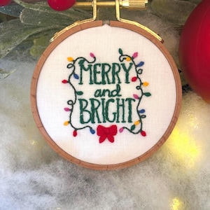May include: Embroidered Christmas ornament in a wooden hoop. The white fabric features the words "MERRY and BRIGHT" in green, surrounded by colorful string lights and a red bow. A festive holiday decoration.