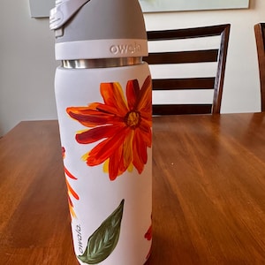 Puede incluir: Botella de agua Owala blanca con tapa gris y banda de acero inoxidable. La botella est&aacute; decorada con grandes flores naranjas y amarillas y hojas verdes. La palabra "owala" est&aacute; impresa en la botella.