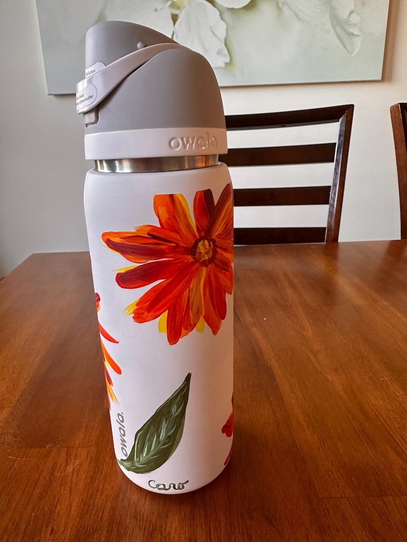 Puede incluir: Botella de agua Owala blanca con tapa gris y banda de acero inoxidable. La botella est&aacute; decorada con grandes flores naranjas y amarillas y hojas verdes. La palabra "owala" est&aacute; impresa en la botella.